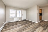 This single-floor unit features a practical, well-organized layout visible in its open floor plan. The living area greets occupants at the front entry, where a white front door opens to a space finished with wood-style luxury vinyl plank flooring in warm neutral tones. Taupe-painted walls and a textured ceiling run throughout the main living spaces, creating a cohesive aesthetic. Natural light enters through multiple windows, and a wall-mounted HVAC unit alongside baseboard heating provides climate control throughout.

The galley-style kitchen is efficiently designed with warm oak cabinetry, dark speckled countertops, a stainless steel sink, and a dark tile backsplash. Black appliances, including a refrigerator and range, complete the kitchen. A window at the end of the kitchen brings in additional natural light.

The bedroom features neutral carpet, dual windows overlooking neighboring structures, a sliding wood-panel closet door, and a ceiling fan. Baseboard heating continues in this room as well.

The full bathroom includes an oak vanity with a dark countertop and single basin sink, a tub-shower combination with a white surround, a small window for ventilation, and wood-style flooring consistent with the rest of the unit.