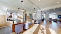 This thoughtfully designed single-floor space offers a generous and open layout that seamlessly blends functionality with contemporary style. The reception area greets visitors with a warm wood-toned front desk, elegant pendant lighting, and comfortable upholstered seating, creating a professional yet inviting first impression. Light wood-look flooring flows throughout the entire space, unifying each zone and enhancing the sense of openness.

The kitchen and bar area is a standout feature, anchored by a large island with a crisp white quartz countertop and a bold blue base, surrounded by modern bar stools. Matte black pendant lights hang overhead, while a textured gray brick accent wall provides a striking industrial backdrop to dark cabinetry and stainless steel appliances.

Wide hallways display a curated gallery of large-scale abstract artwork in cool blue and neutral tones, adding a creative and polished atmosphere throughout. Soaring ceilings amplify the sense of volume, and a lounge area with bold teal accent walls, open shelving, and plush seating offers a relaxed gathering space. Vertically ribbed wall panels and large-format artwork further elevate the interior's design sophistication. Natural light filters generously through the space, contributing to a bright, airy, and welcoming ambiance overall.