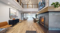 Located at 10805 173rd St W in Lakeville, MN, this well-appointed lobby offers a welcoming and functional communal space spanning a generously sized single floor. The design blends contemporary and transitional styles, highlighted by wide-plank wood-look flooring that runs throughout the entire space, creating a cohesive and warm aesthetic.

Upon entry through large glass-panel doors, visitors are greeted by a dramatic double-height ceiling accented with exposed wood beam details and a statement chandelier overhead. A mezzanine-level overlook with sleek metal railing adds architectural depth and visual interest. Adjacent to the entry is a cozy lounge area anchored by a floor-to-ceiling fireplace topped with a large flat-screen display, flanked by comfortable seating.

Moving deeper into the lobby, distinct zones offer a variety of uses. A casual seating area features plush chairs and a round coffee table set against soft sage-toned walls, while a dedicated dining nook with a marble-top table and upholstered banquette seating provides a more intimate setting. Natural light pours in through large windows and a glass exterior door at the far end.

The space also includes elevator access with stainless steel doors set against a muted teal accent wall, wall sconces, and recessed lighting throughout—ensuring the lobby is both functional and visually refined at all hours.