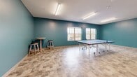 This game room in Madison, WI, offers a well-appointed recreational space designed for active social engagement. The single-floor layout provides a generous open area that comfortably accommodates multiple game stations simultaneously, including a foosball table, an air hockey table, and a full-size ping-pong table, allowing several activities to take place without crowding.

The room's design is defined by its bold teal-painted walls, which lend a vibrant, energetic character to the space. Durable stone-look tile flooring in warm, neutral tones runs throughout, providing a practical and attractive surface suited to high-traffic recreational use. Overhead fluorescent linear light fixtures ensure consistent, bright illumination across the entire room.

Natural light enters through multiple windows along one wall, offering views to the exterior and contributing to a lively, open atmosphere. High-top tables with bar stools are positioned near the windows, providing casual seating for spectators or those taking a break between games. A standard entry door provides access to the adjoining areas of the building. Overall, the space is clean, functional, and thoughtfully arranged to serve as an engaging recreational amenity.