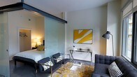 This single-floor space showcases a sleek, contemporary design with an open-concept layout that maximizes both functionality and style. A standout architectural feature is the frosted glass sliding partition, framed in dark metal hardware, which elegantly divides the living area from the sleeping quarters while maintaining a sense of openness and allowing light to flow between the two spaces. The living area is furnished with charcoal-toned seating, including a sofa and accent chairs with yellow-patterned throw pillows, arranged around a glass-top coffee table. An abstract yellow and white wall painting adds a vibrant focal point, complemented by a sleek glass console table beneath it. The sleeping area, visible through the glass partition, features a well-appointed bed with warm, layered bedding and a bedside lamp that casts a soft, inviting glow. The overall color palette of the space blends cool grays and crisp whites with bold yellow accents, creating a modern and energetic ambiance. Clean wall lines, smooth ceilings, and thoughtful furniture placement contribute to an efficient yet stylish interior that feels curated and well-designed throughout.