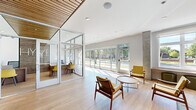 This thoughtfully designed single-floor space combines contemporary architecture with warm, curated interiors to create an inviting and functional environment. The main lobby serves as a welcoming focal point, featuring light hardwood flooring, a striking wood-slat ceiling detail, and an abundance of natural light streaming through expansive floor-to-ceiling windows that frame views of the surrounding greenery and streetscape. Exposed concrete columns add an industrial contrast to the otherwise refined palette of whites and warm neutrals.

The lounge area is anchored by a cognac leather sofa, mid-century modern armchairs in mustard yellow, and pendant lighting with a sculptural quality, lending a relaxed yet polished atmosphere. A glass-walled private office enclosure provides transparency while maintaining acoustic separation, outfitted with a workstation and complementary furnishings.

A standout feature of the space is a bold floral mural in deep navy, cream, and yellow tones, set above a built-in upholstered banquette with marble-topped café tables — creating an intimate and visually dramatic nook. A corridor highlights curated wall art and a solid wood bench, leading to elevator access flanked by exposed concrete and teal-toned doors. Throughout, the design balances artistic expression with practical, welcoming functionality.