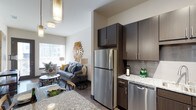 This single-floor unit showcases a contemporary open-concept design that connects the kitchen and living area in one cohesive space. The kitchen is equipped with stainless steel appliances, including a full-size refrigerator and dishwasher, complemented by rich dark wood cabinetry and granite countertops. A subtle tile backsplash adds texture, while pendant lighting above contributes a refined, modern touch.

The kitchen flows directly into the living area, which benefits from large windows that draw in generous natural light, creating a bright and welcoming atmosphere. The living space accommodates comfortable seating and decorative furnishings, with warm-toned hardwood-style flooring running throughout and unifying the two areas visually.

Ceiling height adds a sense of openness to the overall layout. Thoughtful lighting fixtures, including both pendant and overhead options, ensure the space is well-lit throughout the day and evening. The combination of dark cabinetry, light stone countertops, and neutral wall tones creates a balanced, upscale aesthetic throughout this well-appointed single-level residence.