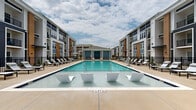 Located at 98 Northwood Blvd in Pittsboro, NC, this two-story residential community showcases a modern architectural design that blends navy blue, warm tan, and crisp white exterior finishes across multiple building wings. The standout feature of the property is its expansive resort-style pool courtyard, which serves as the central gathering space for residents. The large rectangular pool is equipped with in-water lounging chairs, allowing residents to relax directly in the water. The surrounding deck is generously outfitted with chaise lounges, shaded umbrella seating, and dedicated relaxation zones positioned along both sides of the pool. A covered pavilion anchors one end of the courtyard, providing a shaded retreat for outdoor socializing. An additional amenity area extends beyond the pool zone, featuring what appears to be an outdoor kitchen or grilling station, a pergola structure, and a manicured lawn area — ideal for recreation and gatherings. The buildings feature private balconies on upper floors and screened enclosures, offering residents elevated views of the courtyard. The community's cohesive design, well-maintained landscaping, and abundance of outdoor amenities create an inviting, resort-like atmosphere throughout the property.