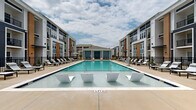 Located at 98 Northwood Blvd in Pittsboro, NC, this two-story residential community showcases a modern architectural design that blends navy blue, warm tan, and crisp white exterior finishes across multiple building wings. The standout feature of the property is its expansive resort-style pool courtyard, which serves as the central gathering space for residents. The large rectangular pool is equipped with in-water lounging chairs, allowing residents to relax directly in the water. The surrounding deck is generously outfitted with chaise lounges, shaded umbrella seating, and dedicated relaxation zones positioned along both sides of the pool. A covered pavilion anchors one end of the courtyard, providing a shaded retreat for outdoor socializing. An additional amenity area extends beyond the pool zone, featuring what appears to be an outdoor kitchen or grilling station, a pergola structure, and a manicured lawn area — ideal for recreation and gatherings. The buildings feature private balconies on upper floors and screened enclosures, offering residents elevated views of the courtyard. The community's cohesive design, well-maintained landscaping, and abundance of outdoor amenities create an inviting, resort-like atmosphere throughout the property.