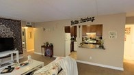 This single-story home showcases an open-concept design that seamlessly connects the living area and kitchen, creating a spacious and functional flow. The living area features neutral-toned carpet and warm beige walls, contributing to an inviting and comfortable atmosphere. A pass-through counter with a granite-style surface bridges the living space and kitchen, enhancing the open feel while maintaining a defined cooking zone. The kitchen is equipped with rich, dark-stained cabinetry, a classic tile backsplash with a brick-like pattern, a white refrigerator, and a stainless-style range, offering both functionality and visual contrast. Tile flooring in the kitchen provides a practical transition from the carpeted living area. A hallway visible on the left suggests additional rooms beyond the main living space. The neutral wall color throughout creates a cohesive backdrop, while white baseboards add a clean, finished look. The layout is well-suited for everyday living, with the kitchen and living areas working together to form a central, communal hub within the home.