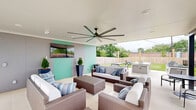 Located at 340 Legion Dr in Lexington, KY, this thoughtfully designed outdoor amenity space blends modern aesthetics with functional entertaining features. The space is anchored by a covered patio lounge area with a crisp white ceiling and a large-span ceiling fan, providing comfortable airflow and shade. The lounge is furnished with wicker sofas and chairs accented by blue and white striped cushions, centered around a coffee table — creating an inviting gathering area. A wall-mounted flat-screen TV is positioned against a two-toned gray and teal accent wall, adding a recreational element to the outdoor experience.

Adjacent to the lounge, an outdoor kitchen island with a built-in gas grill and dark countertop serves as the hub for al fresco dining and cooking. A modern dining table with bench seating sits nearby, all set beneath strands of overhead string lights that extend across wooden posts, lending a warm ambiance during evening hours. A secondary bistro table with chairs provides an additional intimate seating option.

The space opens to a well-maintained, fenced green lawn, offering open space for recreation. The building exterior features a contemporary gray and teal paneled façade with large windows. The combination of covered and open-air zones makes this a versatile, year-round outdoor destination.