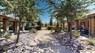Located at 240 Desert Pass St in El Paso, Texas, this single-level outdoor community space masterfully blends Southwestern architecture with thoughtfully designed desert landscaping. Broad paver walkways wind through the grounds, flanked by a rich variety of native and adaptive plantings, including agave, prickly pear cactus, ornamental grasses, and vibrant flowering shrubs, all accented by decorative river rock and boulders that evoke the natural beauty of the Chihuahuan Desert.

Mature shade trees line the pathways and provide generous canopy coverage throughout, creating a cool, inviting atmosphere even under the bright West Texas sun. The grounds are punctuated by a covered pergola structure supported by stacked-stone columns with a pitched roof, offering a shaded outdoor gathering area complete with seating — ideal for relaxation or community events.

Well-maintained green lawn areas contrast with the xeriscaped desert beds, while wrought iron fencing with gated entry points establishes a sense of security and definition throughout the property. Architectural detailing on the surrounding buildings — including stacked-stone facades and warm earth-tone stucco — complements the overall desert-contemporary aesthetic. The outdoor fitness area and multiple seating zones further enhance the functionality and appeal of this inviting community space.