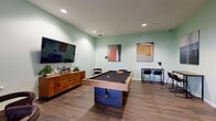 Located at 73 Brownstone Way in Englewood, NJ, this well-appointed community game lounge offers a comfortable and social atmosphere designed for recreation and leisure. The single-floor space is finished with warm wood-look tile flooring and soft sage-green walls that lend a calm, welcoming ambiance throughout.

At the center of the room stands a full-size pool table, serving as the focal point of the space. Adjacent to it is a dedicated poker table surrounded by cushioned chairs, perfect for game nights. A wall-mounted flat-screen TV is paired with a warm wood credenza along one wall, creating an entertainment corner ideal for watching sports or gaming. Multiple bar-height tables with modern stools provide additional seating for socializing or casual dining.

Contemporary abstract artwork adorns the walls, adding a curated aesthetic to the space. Recessed ceiling lighting ensures the room is well-lit throughout. A natural window brings in daylight and offers a view of the surrounding exterior. Glass-paneled doors connect this lounge to adjacent common areas, maintaining an open and integrated feel within the broader community.

The overall design balances a modern, clean aesthetic with comfortable, functional furnishings, making this lounge an inviting destination for residents seeking entertainment and social connection.