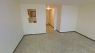 This single-floor space offers a clean, functional layout centered around a carpeted living area finished in neutral tones. The room is framed by white walls and dark baseboards, creating a simple, understated aesthetic. A rectangular pass-through window cut into one wall connects the living area to the kitchen, where wood-toned cabinetry and a sink are visible, allowing for an open sightline between the two spaces without fully removing the wall. The kitchen features traditional wood cabinetry and appears compact yet practical. From the living area, a hallway extends toward additional rooms, illuminated by a ceiling-mounted light fixture that provides consistent overhead lighting. The hallway's white walls and continued carpeting maintain a cohesive look throughout. The overall ambiance is modest and move-in ready, with a neutral color palette that lends itself well to a variety of furnishing styles. The layout is straightforward and efficient, making good use of the available floor space across a single level.