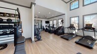 This fitness center is designed to support a comprehensive workout experience across several distinct zones. The main exercise floor is an open, airy space characterized by soaring ceilings, recessed lighting, and light wood-style flooring that runs throughout. Large gray-trimmed windows line the walls, flooding the interior with natural light and offering views of the surrounding exterior. A ceiling fan overhead aids in air circulation during workouts.

The cardio area is well-stocked with a lineup of True-brand treadmills, ellipticals, stair climbers, and a rowing machine, several of which are positioned beneath a wall-mounted flat-screen television for entertainment during exercise. Adjacent to this zone, the strength training area features a full dumbbell rack with a wide range of weights, a multi-functional cable machine, a plate-loaded weight rack, and adjustable benches for varied resistance training.

A separate, enclosed spin room provides a focused cycling environment equipped with multiple stationary bikes and its own wall-mounted television. The glass-paneled entry doors connecting the main fitness floor create a sense of openness while maintaining distinct workout zones throughout this thoughtfully organized facility.