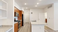 This single-floor residence offers a clean, contemporary design with a cohesive aesthetic carried throughout each space. The kitchen serves as a central highlight, featuring warm wood cabinetry that extends to the ceiling, granite-speckled countertops, and a full suite of stainless steel appliances including a gas range, microwave, and refrigerator. A peninsula with an integrated sink and matching granite surface provides additional prep space and separates the kitchen from the open living area, which is bathed in natural light from large windows fitted with blinds. Recessed lighting throughout the kitchen and living areas adds a bright, polished finish.

Light wood-style flooring runs continuously through the main living spaces, enhancing the sense of openness. The bathroom features a wood vanity with a granite countertop, an integrated sink, a large wall mirror, and an elegant pendant light fixture. A dedicated laundry room houses a full-size washer and dryer with overhead shelving for storage. The spacious walk-in closet is outfitted with wire shelving and hanging rods, offering generous organization options. The overall ambiance is bright, functional, and well-appointed.