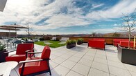 Located at 10 Campus View Ct in Poughkeepsie, New York, this contemporary rooftop terrace offers an exceptional outdoor experience defined by thoughtful design and sweeping panoramic views of the Hudson River and surrounding Hudson Valley mountains. The terrace is finished with large-format light granite-toned pavers that extend across the generous open-air deck, providing a clean, polished aesthetic throughout. A vibrant green turf lawn area introduces a natural element to the rooftop, offering a versatile space for relaxation or casual gatherings. Multiple distinct seating zones are arranged across the terrace, furnished with bold red sofas and chairs alongside neutral lounge pieces, accommodating both intimate conversation areas and larger group settings. Substantial dark planters filled with ornamental grasses and young trees add greenery and visual structure to the layout. A covered indoor-outdoor pavilion anchors one end of the terrace, clad in warm wood paneling beneath a sleek flat roof with recessed lighting, and features large glass windows and a wall-mounted screen, extending the usability of the space in varied weather. A shaded dining area with a striped umbrella provides an additional gathering spot. The terrace benefits from open skies and unobstructed sightlines, making it an ideal space for both daytime enjoyment and evening events.