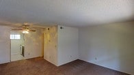 This single-floor residential space presents a straightforward and functional layout designed for comfortable everyday living. The main living area is finished with wall-to-wall carpeting in a neutral tone, providing a warm and understated foundation throughout the space. Crisp white walls keep the interior feeling clean and open, offering a versatile backdrop that can easily accommodate a variety of furnishings and décor styles. A textured popcorn ceiling runs throughout, adding a classic residential character to the space. A ceiling fan is installed in the living area, contributing to year-round comfort and air circulation. The HVAC system is served by ceiling-mounted vents, ensuring efficient climate control throughout the interior. The layout features a defined partial wall that helps delineate areas within the open floor plan, adding a sense of structure without compromising the overall flow. Electrical outlets and light switches are conveniently positioned along the walls, reflecting a practical and accessible design. The overall ambiance is calm and neutral, making this space a solid canvas ready for personalization and everyday use.