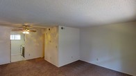 This single-floor residential space presents a straightforward and functional layout designed for comfortable everyday living. The main living area is finished with wall-to-wall carpeting in a neutral tone, providing a warm and understated foundation throughout the space. Crisp white walls keep the interior feeling clean and open, offering a versatile backdrop that can easily accommodate a variety of furnishings and décor styles. A textured popcorn ceiling runs throughout, adding a classic residential character to the space. A ceiling fan is installed in the living area, contributing to year-round comfort and air circulation. The HVAC system is served by ceiling-mounted vents, ensuring efficient climate control throughout the interior. The layout features a defined partial wall that helps delineate areas within the open floor plan, adding a sense of structure without compromising the overall flow. Electrical outlets and light switches are conveniently positioned along the walls, reflecting a practical and accessible design. The overall ambiance is calm and neutral, making this space a solid canvas ready for personalization and everyday use.