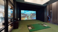 This single-floor entertainment space is purpose-built around a state-of-the-art Golfzon Vision Plus indoor golf simulator. The simulator bay is enclosed with dark-painted walls and features a large high-definition projection screen, a synthetic turf hitting mat with an integrated tee station, and professional-grade tracking technology, creating an immersive virtual golfing experience. Glass-panel partitions separate the simulator bay from the adjoining lounge, allowing clear sightlines between the two areas.

The lounge area is designed for comfortable viewing and socializing. It features a generously sized leather sectional sofa paired with a wood coffee table, anchored by a large-format wall-mounted television and a ceiling-mounted projector. A striking organic wood sculpture adorns one wall, adding an artistic focal point. Floor-to-ceiling draped curtains line the walls, contributing to the room's refined, club-like ambiance and acoustic quality. A built-in wet bar with a bold stone countertop and bar seating runs along one side of the lounge, complemented by recessed lighting overhead.

Dark, large-format tile flooring transitions seamlessly into the green turf of the simulator area. A golf bag storage rack is conveniently positioned near the simulator entrance, completing this well-appointed, turn-key entertainment suite.