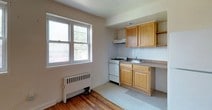 This single-floor unit offers a straightforward, functional layout highlighted by warm hardwood flooring that extends throughout the main living areas, lending a cohesive and inviting feel to the space. The open living area is generously proportioned, featuring clean white walls, natural light from a window, and classic hardwood floors that add character and warmth. A radiator provides in-unit heating, reflecting the building's traditional construction.

The kitchen is efficiently designed, tucked into a dedicated alcove that connects seamlessly to the living space. It features honey-toned wood cabinetry with open shelving, a white gas range, a full-size refrigerator, a sink with a countertop, and a white tile backsplash. A double window allows natural light to fill the kitchen area, brightening the functional workspace.

The bathroom showcases a distinctive retro aesthetic, with bold mint-green ceramic tiles accented by black border trim covering the walls of the tub surround. The bathtub and shower combination is fitted with a handheld showerhead, while a window above provides ventilation and natural light. A decorative curtain adds a personalized touch to the space, completing the unit's blend of classic character and practical design.