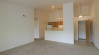 This single-story space features an open-concept design that connects the living area to the kitchen through a raised breakfast bar peninsula. The living area is finished with wall-to-wall neutral carpet, contributing to a warm and understated aesthetic. White walls throughout the space create a clean, bright backdrop, while vaulted ceilings add a sense of vertical openness that enhances the overall feel of the interior.

The kitchen is partially enclosed by a half-wall with a raised bar-height counter, offering both visual separation and a casual dining or serving surface. Inside the kitchen, warm-toned wooden cabinetry provides storage along the upper walls, complemented by white appliances including a refrigerator and range. A small chandelier hangs above the kitchen area, adding a touch of decorative detail to the functional space.

A hallway visible to the left of the living area leads to additional rooms, suggesting a thoughtfully segmented floor plan. The HVAC vent visible on the upper wall indicates a centralized climate control system. Overall, the space presents a functional and straightforward layout with classic finishes throughout.