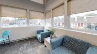 Located at 44 Salix Ct in Baltimore, MD, this single-story commercial office space combines modern design with a bright, functional layout. The open-plan work area is defined by impressively tall ceilings with clerestory windows that flood the interior with natural light, complemented by sleek linear pendant lighting suspended overhead. Exposed ductwork adds an industrial touch to the otherwise polished aesthetic.

The main workspace features multiple workstations with contemporary furniture, including a large executive desk with ergonomic seating and a dedicated collaboration area with a round table and vibrant orange chairs. A built-in service counter with warm wood cabinetry anchors the reception zone, which is further enhanced by a wall-mounted display screen. Patterned carpet tile flooring throughout the office area adds texture and visual interest.

A cozy lounge nook, lined with double rows of windows and fitted roller shades, provides a comfortable retreat with cushioned blue accent chairs and views of the surrounding neighborhood. The space also includes a well-appointed galley-style break room with white shaker cabinetry, stone countertops, stainless steel appliances, a lime green tile backsplash accent, and wood-look flooring. The overall design is clean, contemporary, and highly functional, making it well-suited for a professional office environment.