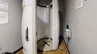 This compact, single-room tanning space at 100 Hawk Ridge Dr in Iowa City, Iowa, is purpose-built for UV tanning services. The room is efficiently laid out to accommodate a full stand-up cylindrical tanning bed as its centerpiece, equipped with vertical UV lamps and a marked standing platform. The tanning unit is flanked by a wall-mounted control panel, providing straightforward operation management for the session.

The surrounding space is functional and neatly organized. A wall-mounted sanitation dispenser is positioned for easy access, accompanied by posted hygiene instructions reminding users to wipe down the tanning bed after use. A wooden ladder-style shelving unit offers storage for tanning accessories and personal products, while a wooden chair provides a convenient seating option. Informational and safety notices, including ultraviolet radiation warnings, are clearly displayed on the walls.

The room features neutral gray walls, a solid black entry door with a lever-style handle, and durable flooring. Overhead lighting ensures adequate visibility throughout the space. The overall design is utilitarian and clean, prioritizing safety, hygiene, and ease of use, making it a straightforward and well-equipped individual tanning room suitable for a fitness or wellness facility setting.