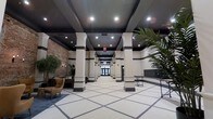 This expansive lobby presents a striking blend of classic and contemporary design elements. The space is defined by a long, symmetrical corridor flanked by tall, cream-colored columns with dark accent banding, lending a formal and elegant architectural character. The columns extend toward glass entry doors at the far end, creating a strong sense of depth and grandeur.

The flooring features a bold geometric tile pattern in cream and dark tones, adding visual interest while reinforcing the space's refined aesthetic. Overhead, a dark-painted ceiling with recessed lighting provides a dramatic contrast to the lighter walls and columns, ensuring the corridor remains well-illuminated throughout.

To one side, an exposed brick wall introduces a raw, historical texture that complements the otherwise polished finishes, providing a thoughtful juxtaposition of old and new. This area is furnished with a seating arrangement that includes a tan leather armchair, offering a comfortable lounge vignette.

On the opposite side, a built-in mailbox bank and a slim console table serve as practical amenities for residents. Lush tropical plants are strategically placed throughout, adding warmth and a refreshing natural element to the otherwise structured and formal environment.