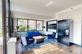 The Bartlett Court Apartments lobby in Streamwood, Illinois, serves as the welcoming gateway to the community and is thoughtfully designed to be both functional and inviting. The space features wood-look tile flooring throughout, lending warmth and a modern aesthetic that flows seamlessly across the entire area.

The main lounge area is furnished with a coordinated set of rich royal blue velvet seating — including a sofa and accent chairs — arranged around a solid wood coffee table atop a patterned area rug. Large black-framed windows wrap the lounge, flooding the space with natural light and offering views of the surrounding landscaping and grounds. A prominent Bartlett Court Apartments branded sign anchors the feature wall, reinforcing the community's identity.

Adjacent to the lounge, a bank of dark navy smart package lockers provides residents with a secure and convenient solution for parcel delivery. A separate glass-enclosed leasing office is visible from the main lobby, accessible via a black-framed glass partition wall and door, with a reception desk and full kitchen amenity visible inside.

A classic wooden bench near the entryway offers additional seating, while the entry corridor features a recessed mat and easy access to the rest of the building. The overall ambiance is clean, bright, and professionally appointed, creating a polished first impression for residents and visitors alike.