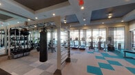 Located at 5330 Sienna Pkwy in Missouri City, TX, this single-floor fitness center is designed to support a comprehensive workout experience. The space features a modern aesthetic defined by coffered ceilings with recessed lighting, a bold teal and neutral checkered carpet pattern, and a striped accent wall that adds visual depth and energy to the environment. Large floor-to-ceiling windows along one side of the facility flood the space with natural light and provide outdoor views, enhancing the open and airy atmosphere. Full-length mirrors are strategically placed throughout to maximize the sense of space and assist with form during exercise. The facility is divided into functional zones: a cardio area lined with treadmills and elliptical machines, a strength training section housing a tiered dumbbell rack with a wide range of weights, and a functional training area equipped with a cable and pulley multi-station system, a punching bag, and a wall-mounted RAX training rig with suspension straps. Additional selectorized strength machines, including a Matrix leg press unit, round out the equipment offerings. A wall-mounted flat-screen TV is positioned for visibility from the free weights area. The entrance features a glass door with decorative patterned wall panels, offering a polished and welcoming entry point into the space.