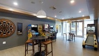 This single-floor amenity space is designed as a lively entertainment and social hub, combining retro gaming culture with a modern, welcoming atmosphere. The room features a collection of classic upright arcade cabinets and pinball machines, including iconic titles, positioned along one wall and serving as the focal point of the space. Overhead, stylish white pendant lights hang from a clean, recessed-lit ceiling, casting warm, even illumination throughout. The walls are painted in a rich slate blue with warm accent trim, lending the space a contemporary yet relaxed character. Round high-top tables paired with wooden bar stools are scattered across the light-toned floor, offering casual seating for groups to gather and socialize. Toward the rear of the space, large windows and glass doors allow an abundance of natural light to filter in, connecting the interior to the outdoors and creating an open, airy feel. The overall layout is open and unobstructed, making the space feel spacious and easy to navigate. This well-appointed game lounge is thoughtfully designed to encourage interaction and leisure, making it an ideal communal amenity for residents or guests seeking entertainment and a sense of community.