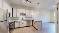 This thoughtfully designed single-floor unit offers a clean, contemporary aesthetic with an open-concept layout that seamlessly connects the kitchen and living areas. The kitchen is a standout feature, outfitted with light wood-toned cabinetry accented by matte black hardware, speckled granite countertops, and a full suite of stainless steel appliances including a refrigerator, electric range, microwave, and dishwasher. A peninsula with matching granite provides additional counter space and serves as a natural dividing point between the kitchen and the expansive living area. Stylish pendant lights and a track lighting system illuminate the kitchen, adding a modern, layered lighting effect. Light wood-style plank flooring runs throughout the unit, contributing to a bright and cohesive feel. The living space is generously sized and bathed in natural light from windows dressed with clean white blinds. The bathroom features the same consistent design language, with granite-topped white cabinetry, a tub-shower combination, and wood-style flooring. The entry area provides practical access to additional interior doors, completing a well-organized and efficiently laid out living space.
