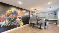 Located in Mill Creek, WA, this professionally designed pet grooming studio is a compact yet highly functional space that makes a striking first impression. The most commanding feature is a large, colorful hand-painted dog mural set against a dramatic black accent wall, immediately establishing the space's playful and pet-focused identity. Light wood-look flooring runs throughout, complementing the soft blue-gray walls that define the rest of the interior.

At the heart of the grooming area stands a stainless steel elevated bathing tub with step access, positioned beneath an industrial-style pendant light fixture with exposed Edison bulbs suspended from a wooden beam. Adjacent to the tub is a modern cabinetry island with a white countertop and integrated sink, providing a clean and practical workspace. A wall-mounted professional pet dryer and a dedicated grooming table with built-in workspace complete the service setup.

Natural light enters through multiple windows, offering views of the surrounding greenery and keeping the space feeling open and airy. Wall-mounted wire baskets, treat dispensers, and framed pet photography add functional and decorative charm throughout. A "Wipe Your Paws" entry mat at the main door adds a welcoming touch. The studio is efficiently organized to support a full-service grooming workflow within a cozy, well-appointed footprint.