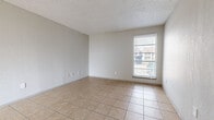 This single-story space is designed with a straightforward, functional layout evident from the 3D floor plan, which reveals a kitchen, living area, dining nook, bathroom, and additional rooms connected by interior doorways. Consistent light beige ceramic tile flooring runs throughout the unit, providing a cohesive and easy-to-maintain surface. The walls are painted in neutral light gray tones, complemented by textured popcorn ceilings that are characteristic of the unit's construction style.

The living area is generously proportioned, featuring a ceiling fan, a ventilation duct, and access to what appears to be a patio via vertical blinds, allowing for natural light. A separate, windowed room adjacent to the kitchen serves well as a dining space, with a tall window fitted with horizontal blinds offering outdoor views.

The kitchen is equipped with white appliances including a refrigerator, electric range, and dishwasher, alongside gray cabinetry, speckled laminate countertops, and a double stainless steel sink. The bathroom includes a white tiled tub and shower surround. Throughout, multiple windows ensure natural light reaches the interior, contributing to a bright and open ambiance.