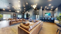 Located at Reflection Pointe in Hamilton, Ohio, this single-floor space offers a generously sized, open-concept living area designed with a distinctly upscale aesthetic. The interior is defined by its dramatic vaulted ceilings finished in a deep, textured navy-blue tone, complemented by coordinating wainscoted walls that lend a refined architectural character throughout. Rich hardwood flooring runs the length of the space, anchoring the various seating zones with warmth and continuity.

The living room serves as the centerpiece, featuring a wood-burning fireplace with a rustic wood mantel set beneath a wall-mounted display screen, flanked by elegant panel molding. Multiple seating arrangements — including cognac leather sofas, navy velvet accent chairs, and plush swivel lounge seats — create distinct zones for conversation and relaxation. A sculptural globe chandelier provides a striking focal point overhead, supplemented by recessed lighting throughout.

An arched doorway connects the main living area to a secondary lounge zone and a built-in bar or coffee station, adding functional versatility. Large windows along multiple walls allow natural light to filter in, softening the bold color palette. A bar counter with high stools offers an additional gathering space. The overall ambiance is contemporary yet inviting, blending bold design choices with comfortable, well-appointed furnishings.