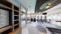 This single-floor fitness center is thoughtfully designed to deliver both functionality and visual appeal. The space is divided into distinct exercise zones, accommodating a wide range of cardio equipment including Matrix treadmills, elliptical trainers, stair climbers, and step mills. Large windows line the walls, flooding the interior with natural light and providing outdoor views that enhance the workout experience.

A standout architectural feature is the dark slatted ceiling, which adds a bold, contemporary contrast to the light-toned walls painted in soft blue and white hues. Ceiling fans are mounted throughout, ensuring proper air circulation. Recessed lighting supplements the natural light for a consistently bright environment.

The flooring features an eye-catching geometric hexagonal tile pattern in neutral tones of grey, beige, and white, adding a modern design element underfoot. Full-length mirrors are strategically placed to visually expand the space and allow users to monitor their form. Built-in rustic wooden open shelving provides convenient storage.

Wall-mounted flat-screen televisions are positioned throughout the space for entertainment during workouts. Glass doors and partitions define separate workout zones while maintaining an open, connected feel throughout the fitness center.
