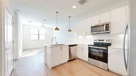 This thoughtfully designed single-level residence offers a clean, modern aesthetic throughout its one bedroom and one bathroom layout. The open-concept kitchen serves as the centerpiece of the living area, featuring crisp white shaker cabinetry, a peninsula with an undermount sink, light quartz countertops, and a full suite of stainless steel appliances including a range, over-the-range microwave, dishwasher, and French door refrigerator. Two statement gold pendant lights hang above the peninsula, adding a warm, contemporary accent. Light wood-look flooring runs through the kitchen and living areas, lending the space a bright and cohesive feel. A ceiling fan provides additional comfort in the living room, which benefits from natural light through a window.

The bedroom is a comfortable and quiet retreat, finished with soft wall-to-wall carpeting and a window with blinds that allows natural light to filter in. The full bathroom is well-appointed with white cabinetry, a vanity with ample storage, a soaking tub with a shower surround, and wood-look flooring. A dedicated closet accessible from the bathroom provides convenient storage. The overall palette of crisp whites and neutral tones creates a clean, airy ambiance throughout the entire residence.