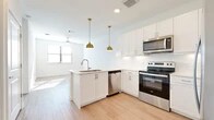 This thoughtfully designed single-level residence offers a clean, modern aesthetic throughout its one bedroom and one bathroom layout. The open-concept kitchen serves as the centerpiece of the living area, featuring crisp white shaker cabinetry, a peninsula with an undermount sink, light quartz countertops, and a full suite of stainless steel appliances including a range, over-the-range microwave, dishwasher, and French door refrigerator. Two statement gold pendant lights hang above the peninsula, adding a warm, contemporary accent. Light wood-look flooring runs through the kitchen and living areas, lending the space a bright and cohesive feel. A ceiling fan provides additional comfort in the living room, which benefits from natural light through a window.

The bedroom is a comfortable and quiet retreat, finished with soft wall-to-wall carpeting and a window with blinds that allows natural light to filter in. The full bathroom is well-appointed with white cabinetry, a vanity with ample storage, a soaking tub with a shower surround, and wood-look flooring. A dedicated closet accessible from the bathroom provides convenient storage. The overall palette of crisp whites and neutral tones creates a clean, airy ambiance throughout the entire residence.