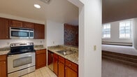 This one-bedroom, one-bathroom residence offers a thoughtful layout across a single floor, combining historic architectural character with functional modern updates. The kitchen is efficiently designed with rich wood cabinetry, granite countertops, a stainless steel sink, and a full suite of appliances including a range and built-in microwave. A pass-through counter connects the kitchen to the living area, enhancing the open feel of the space. The living room is a standout feature, showcasing a dramatic exposed brick accent wall that adds warmth and industrial charm. Carpet flooring runs through the living and sleeping areas, while tile flooring anchors the kitchen. The bedroom benefits from natural light through a large window with views of the surrounding building exterior, soft gray walls, and carpet underfoot. The full bathroom is clean and well-finished, featuring granite countertops at the vanity, a tiled tub-shower combination, and a large mirror with overhead lighting. A hallway connects the main areas of the residence, contributing to a cohesive and efficient flow throughout the home. The exposed brick elements visible from multiple rooms lend the space a distinctive character rooted in its building's history.
