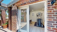 This dedicated fitness center is set within a single-story structure with a classic brick exterior, accessible through glass-panel doors that invite natural light into the space. The interior features clean, white walls and light wood-style flooring that gives the room a bright, open feel, while rubber matting beneath the equipment provides added stability and protection. Recessed ceiling lighting ensures the space is well-lit throughout. The cardio equipment lineup includes elliptical trainers, a stationary recumbent bike, and a treadmill — all from Precor, a reputable commercial fitness brand — catering to a variety of cardiovascular workout preferences. A cushioned workout bench provides a dedicated area for strength and stretching exercises. A wall-mounted flat-screen TV offers entertainment during workouts, and a motivational wall decal reading "Everyday is a Good Day to Work Out" adds an encouraging touch to the atmosphere. Posted fitness area rules and a workout chart ensure users can exercise safely and effectively. The space is compact yet thoughtfully organized, making efficient use of the available floor area to accommodate multiple users simultaneously.