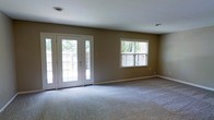 This two-story home offers a thoughtfully designed layout across two floors, combining comfort and functionality. The main living area is generously proportioned, featuring plush wall-to-wall carpeting that adds warmth underfoot. A standout feature of this room is the set of elegant French doors, which flood the space with natural light and provide a seamless connection to the outdoor area beyond. An additional double-hung window further enhances the bright, airy atmosphere, offering views of the surrounding exterior. The walls are finished in a neutral warm taupe tone, creating a versatile backdrop that complements a wide range of interior styles. White trim and baseboards provide a clean, classic contrast throughout. Ceiling-mounted ventilation is integrated discreetly into the space, maintaining a tidy aesthetic. The overall ambiance is open and inviting, with an abundance of natural light lending the interior a fresh, welcoming feel. Spread across two floors, the home provides ample room for both living and private spaces, making it well-suited for a variety of needs and lifestyles.