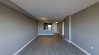 This single-floor unit presents a thoughtfully designed open-concept layout that seamlessly connects the living area to the kitchen. The space is finished with wide-plank, hardwood-style flooring in a rich gray-toned hue that runs throughout, complementing the cool gray walls and crisp white baseboards for a cohesive, modern aesthetic. The textured ceiling adds subtle depth to the interior.

The kitchen is positioned at the rear of the open living space and features dark cabinetry, a stainless steel appliance package, and a raised peninsula that provides both additional counter space and a natural visual divider between the kitchen and the main living area. Track lighting above the peninsula ensures functional task illumination.

A bright entryway is visible adjacent to the kitchen, offering a welcoming transition into the unit. The entry area is well-lit with a ceiling fixture and includes a closet, providing practical storage from the moment you step inside.

The open living area offers generous proportions, allowing for flexible furniture arrangements to suit a variety of lifestyles. The clean lines, neutral tones, and consistent finishes throughout the space create a calm, move-in-ready environment that balances modern design with everyday functionality.