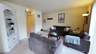This single-floor space features a welcoming living room as its centerpiece, finished with light neutral wall tones and wall-to-wall carpeting that contribute to a warm, understated ambiance. The living area is generously sized, accommodating multiple seating arrangements including a large dark charcoal sofa and a coordinating gray armchair, both oriented around a sleek black coffee table. A soft area rug anchors the seating arrangement, adding texture to the carpeted floor. Decorative wall art, including a framed chandelier illustration, adds a touch of personality to the otherwise clean, minimalist walls. An electrical panel is mounted on one wall, a practical feature integrated discreetly into the space. A flush ceiling light fixture provides overhead illumination throughout the room. The living area connects directly to a hallway corridor, which features a warm wood-toned door and additional access to other areas of the unit. The hallway also includes a small entryway nook, visible with shoe storage, offering a defined transitional zone. Overall, the space presents a functional, comfortable layout with a clean and cohesive aesthetic.