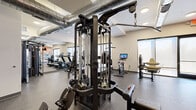 This modern fitness center is designed to support a comprehensive workout experience within a thoughtfully organized, single-floor layout. The space is defined by durable rubber flooring, recessed ceiling lighting, and exposed industrial-style ductwork that lends an urban aesthetic to the interior.

The cardio zone lines one side of the room, featuring a row of treadmills and stationary spin bikes, each equipped with individual digital display screens. Large frosted windows along this wall allow natural light to filter in while maintaining privacy, creating a bright and energizing atmosphere for workouts.

The strength training area houses a multi-station cable machine, a dumbbell rack with a full range of free weights, and dedicated weight benches, providing ample options for resistance training. A wall-mounted mirror expands the visual depth of this zone and allows users to monitor their form during exercises.

A functional training section showcases a storage rack stocked with medicine balls, stability balls, and foam rollers, alongside additional strength machines. This area flows toward a glass door leading to an adjacent room or exterior space, enhancing the sense of openness.

A striking large-scale mural featuring bold, colorful abstract and illustrative artwork serves as a focal point across one full wall, adding a dynamic and motivating visual element to the space. The overall ambiance is clean, modern, and energizing, making this fitness center a well-rounded facility for users of all fitness levels.