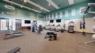 This fitness center in Indianapolis, Indiana, offers a comprehensive workout environment thoughtfully designed for a wide range of training needs. The space features soaring ceilings accented with a striking teal upper wall and crisp white lower walls, giving the room an open, energizing atmosphere. Acoustical panels line the upper walls, contributing to a comfortable sound environment throughout.

The cardio zone is equipped with a row of treadmills, an elliptical, and a stationary cycle, all positioned beneath wall-mounted televisions for an engaging workout experience. A large interior window overlooks an adjacent court, adding visual openness and connectivity to the broader facility.

The strength training area is well-appointed with a cable machine, a Smith machine with bench, a multi-use functional trainer, and a full rack of dumbbells spanning a wide weight range. Free weight benches and a rowing machine provide additional training versatility, while a heavy tire adds a functional fitness element to the floor space.

A dedicated accessory wall features organized open shelving for storage alongside a mirrored section, complementing a rack stocked with stability balls, medicine balls, and kettlebells. Pendant ring lights hang from the ceiling, providing focused illumination across the floor. Durable carpet flooring and rubber mat zones complete this practical, modern fitness facility.