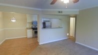 This single-floor residence offers a functional and well-organized open-concept layout that seamlessly connects the living and kitchen areas. The living space features warm, neutral-toned carpeting and is topped with a ceiling fan, providing both comfort and air circulation throughout the room. Crown molding runs along the upper edges of the walls, adding a refined architectural detail to the interior.

The kitchen is well-appointed with stainless steel appliances, including a range with an overhead microwave and a full-size refrigerator. White cabinetry provides ample storage, while the countertops offer generous workspace. A pass-through bar counter connects the kitchen to the living area, creating an interactive and open feel that is ideal for entertaining or casual dining.

The flooring transitions from durable hardwood-style planks in the kitchen to plush carpeting in the living area, clearly defining each functional zone while maintaining a cohesive aesthetic. Visible HVAC vents in the ceiling ensure consistent climate control throughout the space. A hallway visible from the main living area suggests additional rooms beyond, further contributing to the home's practical and livable layout. Overall, the space presents a clean, move-in ready environment with a balance of comfort and functionality.