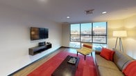 Located at 1120 N Division St in Spokane, Washington, unit 217 is a thoughtfully designed single-floor residence that blends contemporary style with functional living. The open-concept living area is a standout feature, anchored by expansive floor-to-ceiling windows on two sides that flood the space with natural light and offer broad views of the surrounding cityscape. Warm hardwood flooring runs throughout, complementing the clean lines and neutral tones of the interior. A wall-mounted flat-screen television and floating media console add a modern touch to the living space, while the comfortable seating arrangement and bold red accents bring warmth and character to the room. The kitchen is well-appointed with dark espresso cabinetry, stainless steel appliances including a refrigerator, range, and microwave, and a light granite peninsula with bar seating — ideal for both cooking and casual dining. The bedroom offers a serene retreat, featuring plush carpeting with a richly patterned area rug, a king-sized bed with a bold upholstered headboard, and window treatments that allow for privacy and light control. A wall-mounted television adds convenience to the bedroom as well. The bathroom is finished with dark wood cabinetry, a wide vanity with ample counter space, and clean tile flooring, completing this well-curated urban residence.