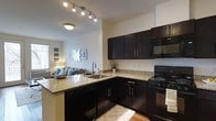 This single-floor residence offers a thoughtfully designed open-concept layout that seamlessly connects the kitchen and living areas, creating a cohesive and welcoming atmosphere. The kitchen is a standout feature, showcasing rich dark cabinetry with sleek hardware that contrasts beautifully against light granite countertops. A peninsula with an undermount sink serves as both a functional workspace and a natural dividing point between the kitchen and living space, ideal for casual dining or entertaining. Black appliances, including a gas range, built-in microwave, and dishwasher, complement the bold cabinetry. Track lighting overhead provides focused illumination throughout the kitchen area, while a flush-mount ceiling fixture adds ambient light. The open living area flows naturally from the kitchen, featuring warm-toned walls and hardwood-style flooring that extends throughout the space. Natural light filters in through windows, brightening the interior and enhancing the overall sense of openness. The living area offers a comfortable, versatile space with room for full furnishings. The combination of modern finishes, functional design, and an open floor plan makes this residence a practical and stylish single-floor home.