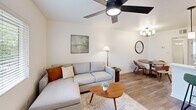 This efficiently designed 1-bedroom, 2-bathroom residence in Phoenix, AZ offers a thoughtfully organized single-floor layout that makes excellent use of its space. The living room serves as the heart of the home, featuring warm wood-look flooring, a sleek ceiling fan with integrated lighting, and a window overlooking the exterior — filling the room with natural light. A sectional sofa, coffee table, and adjacent dining area create a seamless open-concept flow, complemented by a chandelier and decorative accents. The kitchen is visible from the living and dining areas, fitted with stainless steel appliances and light cabinetry. The bedroom is a comfortable, well-lit retreat with wood-look flooring, a large window with blinds, and clean white walls that lend a calm, airy ambiance. Two bathrooms serve the unit: one features a floating white vanity with a square undermount sink and brushed nickel fixtures alongside a closet, while the second includes a full soaking tub and shower combo with large-format cream tile surround and chrome hardware. Throughout, the neutral color palette and consistent flooring create a cohesive, modern aesthetic suited to everyday living.