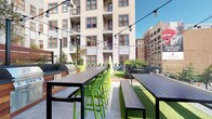 Located at 12 W Gay St in the heart of downtown Columbus, Ohio, this outdoor terrace offers a well-appointed communal gathering space designed for social interaction and urban living. The terrace is defined by its lively, contemporary aesthetic, featuring stretches of artificial turf that provide a refreshing green contrast against the surrounding cityscape.

Multiple long bar-height dining tables paired with bold lime-green chairs create ample seating for groups, while additional bench-style tables offer flexible arrangements for larger gatherings. Overhead, a canopy of string lights spans the length of the terrace, providing an inviting ambiance for both daytime and evening use.

A dedicated outdoor grilling station is available for resident or guest use, adding a functional, social element to the space. Raised planters with greenery line the perimeter, contributing to the terrace's sense of privacy while softening the urban environment.

The open-air layout affords clear views of the surrounding downtown Columbus architecture, including the multi-story residential building above and the historic brick facades of nearby structures. The terrace's single-level design ensures ease of access and flow throughout the space, making it an ideal venue for casual gatherings, outdoor dining, and community events in a dynamic urban setting.