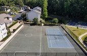 Located at 6915 Roswell Rd in Sandy Springs, Georgia, this outdoor recreational facility features two distinct sport courts set within a lush, tree-lined environment. The primary court is a blue hard-surface tennis court, clearly marked with crisp white boundary lines and equipped with a regulation net, making it suitable for both tennis and pickleball play. The court is fully enclosed by a black chain-link fence, providing a secure and defined playing area. Mature deciduous and evergreen trees surround the perimeter, offering partial shade and a natural backdrop that enhances the outdoor atmosphere.

A secondary hard-surface court is also present on the property, featuring a gray playing surface with white line markings. This court is similarly enclosed and benefits from the same verdant surroundings, along with a brick retaining wall that adds structural definition to its boundary. A children's playground is visible nearby, indicating that this facility is part of a broader recreational amenity area. Ample open sky above both courts ensures excellent natural lighting throughout the day. The overall setting is well-maintained and designed to support active outdoor recreation for users of all skill levels.