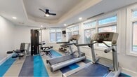 This dedicated fitness center offers a comprehensive workout environment within a thoughtfully designed single-room layout. The space is defined by its distinctive tray ceiling, which adds architectural depth and visual interest, complemented by recessed lighting and a ceiling fan that ensure both comfort and functionality during exercise sessions.

Large, multi-pane windows line the walls, flooding the room with abundant natural light while providing views of the surrounding outdoor greenery. The bright white walls and light-toned ceiling enhance the open, airy atmosphere, making the space feel welcoming and energizing.

The fitness center is equipped with a well-rounded selection of exercise equipment, including multiple treadmills, an elliptical cross-trainer, a stationary bike, and a multi-station cable strength machine with an adjustable bench. A tiered dumbbell rack stocked with a range of free weights supports strength training, while a wall-mounted flat-screen television provides entertainment during workouts. A dedicated coat hook rail near the entrance adds a practical touch.

The floor features a durable striped blue, gray, and white exercise mat surface that defines the workout zones and adds a dynamic visual element to the room. A separate entry door provides access to adjoining facilities. Overall, the space delivers a clean, modern, and fully functional fitness environment suited for a variety of training needs.