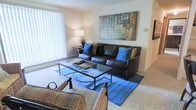 This single-floor residence offers a warm and inviting atmosphere designed for comfortable everyday living. The living room serves as the centerpiece of the space, showcasing plush carpeted flooring and a generous layout that accommodates a full seating arrangement with ease. A large window fitted with vertical blinds floods the room with natural light, creating an airy and open ambiance throughout the day. The seating area features a classic dark leather sofa accented with vibrant blue and tan throw pillows, complemented by a sleek glass-top coffee table that adds a contemporary touch. A bold, colorful abstract canvas artwork anchors the main wall, providing a striking visual focal point. Soft accent lighting from a table lamp adds warmth to the room during evening hours. The neutral wall tones and light-colored carpet create a versatile backdrop that suits a variety of décor styles. Additional seating is thoughtfully incorporated into the layout, making the space well-suited for both relaxed lounging and casual entertaining. Overall, the residence exudes a cozy yet stylish character, balancing functionality with a clean, modern aesthetic.
